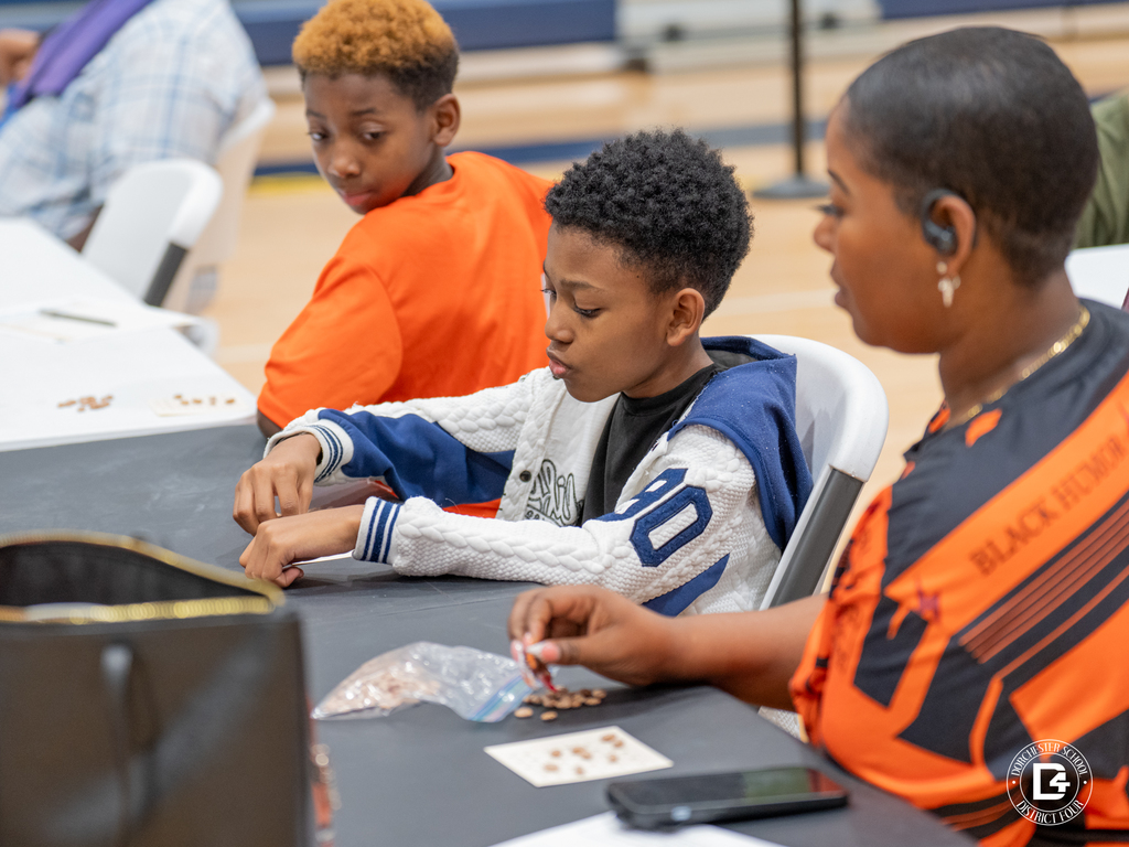 A boy wearing a white jacket focuses on his bingo card, with beans scattered on the table, as his family sits beside him.