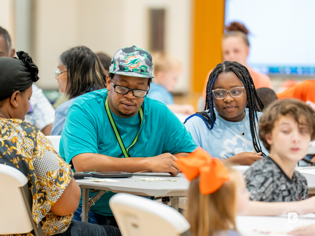 A man in a teal shirt and Miami Dolphins hat sits with a girl and woman as they look over their bingo cards together.