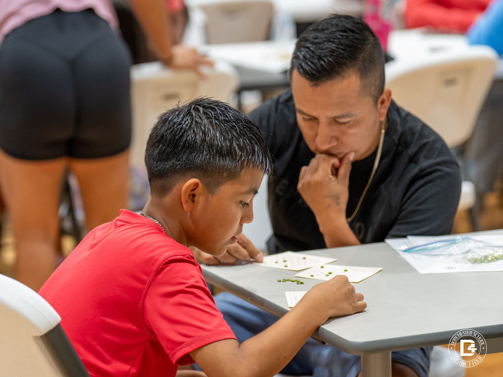 A father and son concentrate on filling out their bingo cards together at a small gray table.