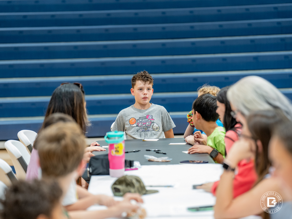 A group of children sit around a long black table playing bingo, one boy in the center looking toward the caller.