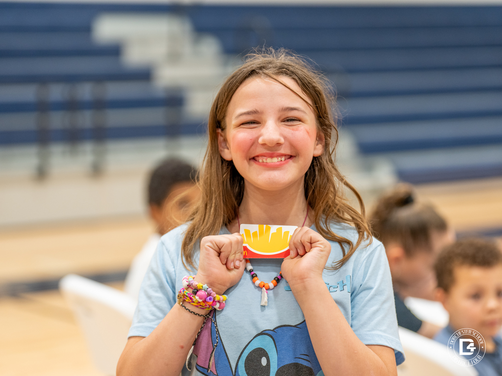 A smiling girl proudly holds up her prize, a gift card, while wearing a light blue shirt and colorful beaded bracelets.
