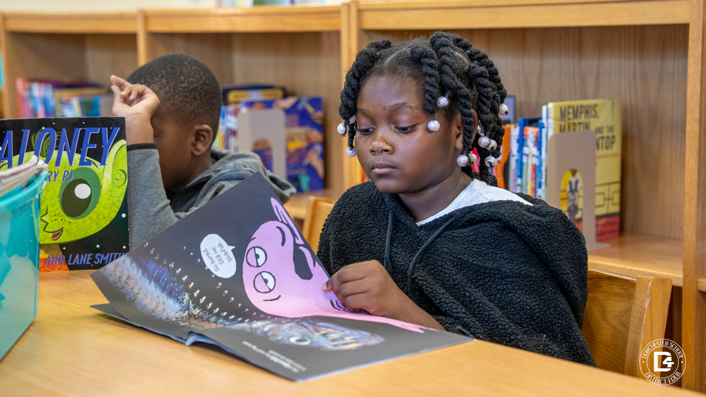 A student sits quietly at a library table, reading an illustrated book titled “Baloney” with focus and interest.