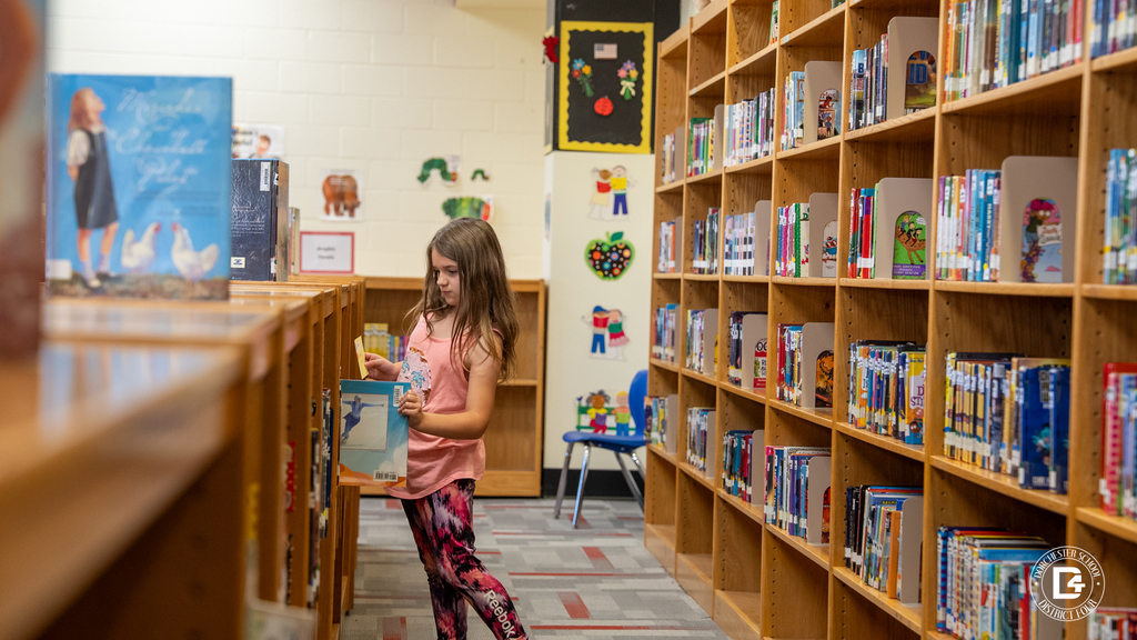 A young girl browses through a bookshelf in the school library, holding a book as she decides what to read next. Colorful decorations and rows of books line the background.