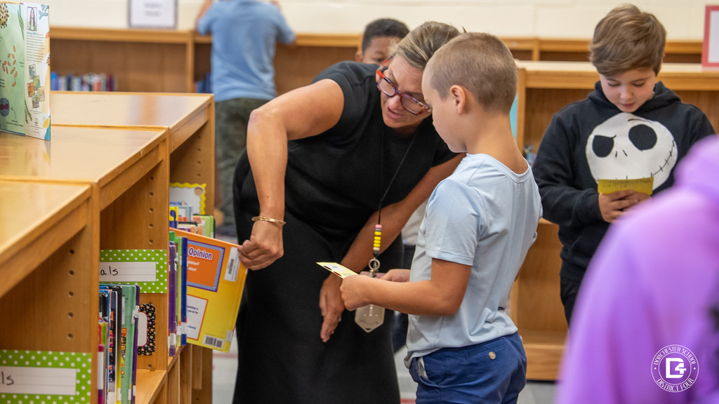 Mrs. Davis helps a student locate a book that matches his reading level while another student nearby checks his list. Library shelves filled with books surround them.