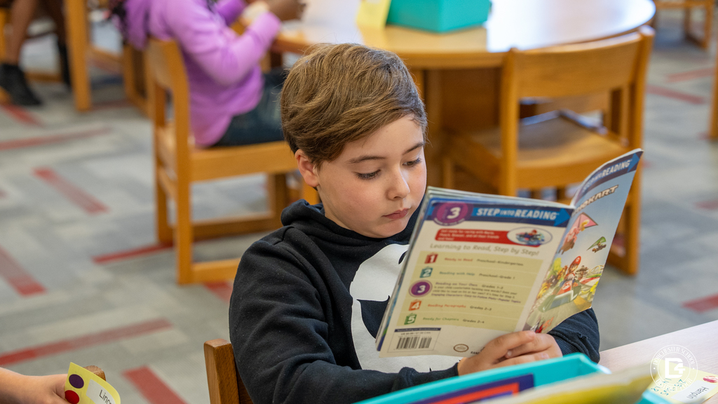 A student in a black hoodie with a skeleton face design reads a “Step into Reading” book at a table in the library.