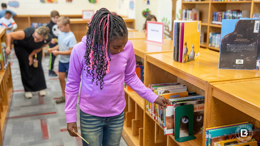 A student wearing a purple hoodie looks through a shelf of library books while Mrs. Davis assists other students in the background.