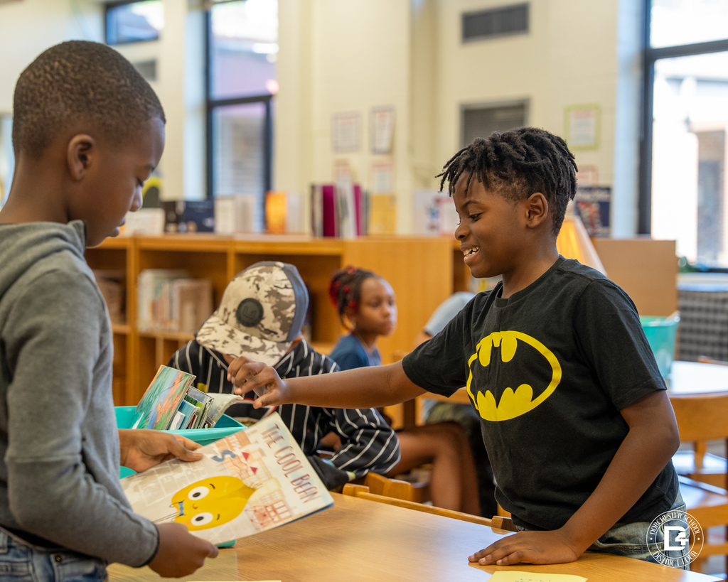Two students look at picture books together at a library table, smiling as they share their selections.