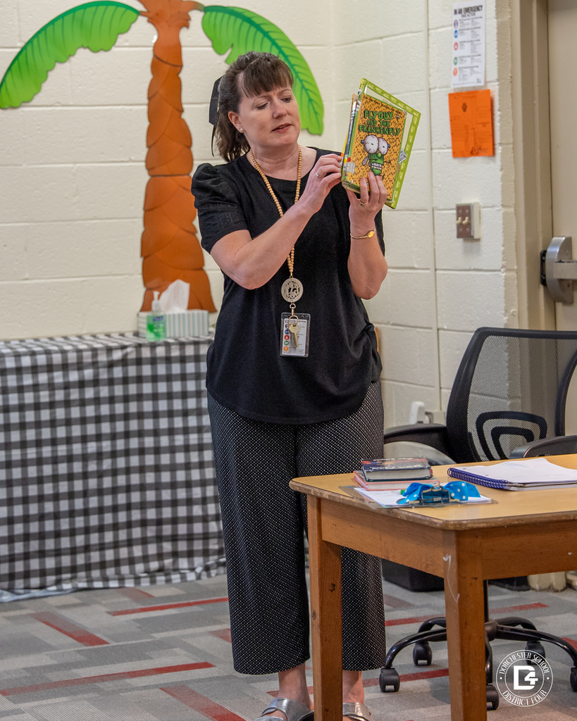 School librarian Mrs. Stephenson stands at the front of the library holding a “Fly Guy” book, explaining something to students seated nearby.