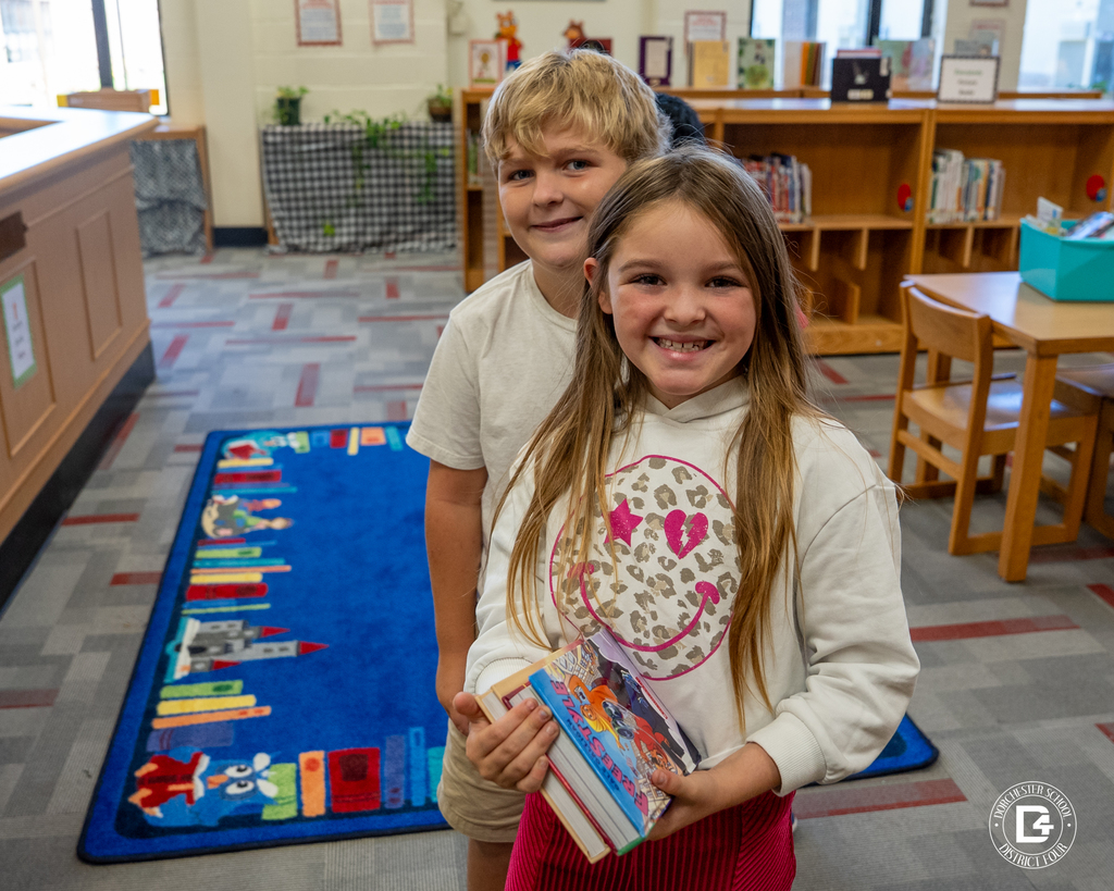 Two smiling students stand side by side in the library, holding a stack of books they’ve selected to check out.