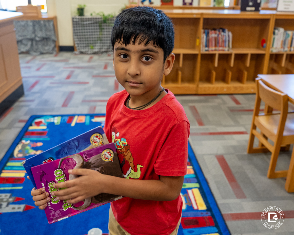 A student in a red shirt holds several books against his chest while standing on a colorful reading rug in the library.