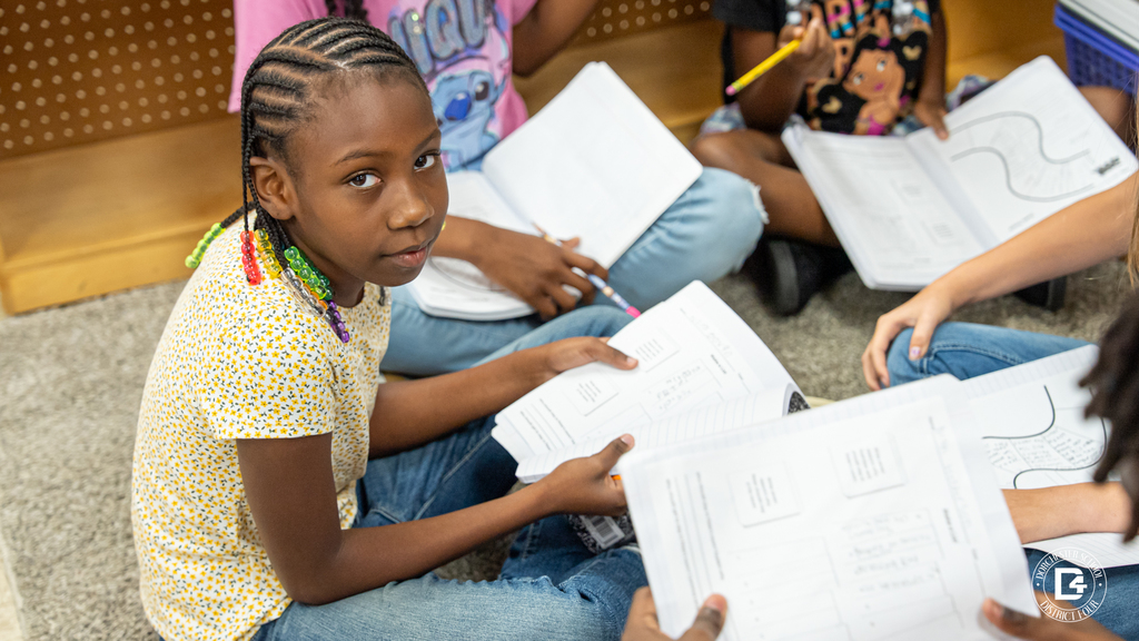 A student sitting on the classroom carpet looks up from her notebook while holding a pencil during a group reading discussion activity. Other students around her are engaged with their workbooks.