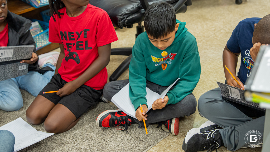 Two students sitting on the floor take notes in their composition books during a small group discussion. They appear focused and engaged in their writing task.