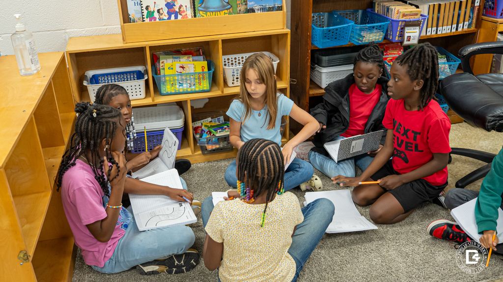 A full group of 4th-grade students sit in a discussion circle on the carpet, each with a notebook open as they share ideas about a story during a compare-and-contrast exercise.