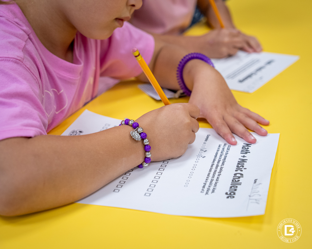 Close-up of a child’s hand holding a pencil and marking answers on the Math + Music Challenge worksheet.