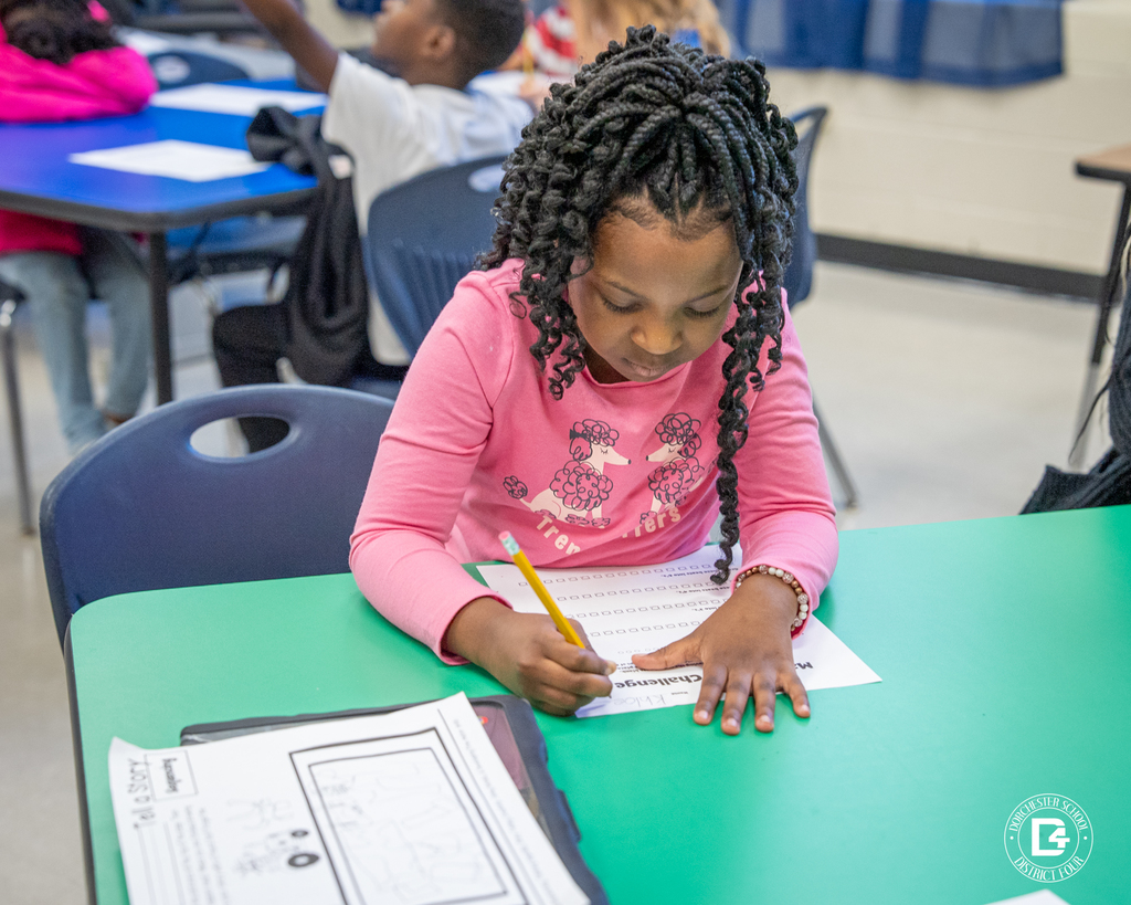 A student in a pink shirt concentrates on her worksheet, writing answers for the Math + Music Challenge at a green table.