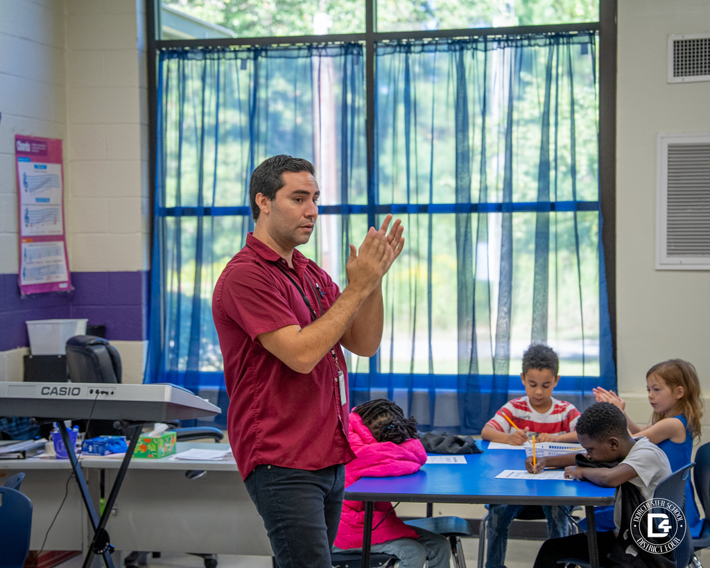 Music teacher Mr. Mansilla claps to a rhythm at the front of the classroom while students listen and work at their tables.