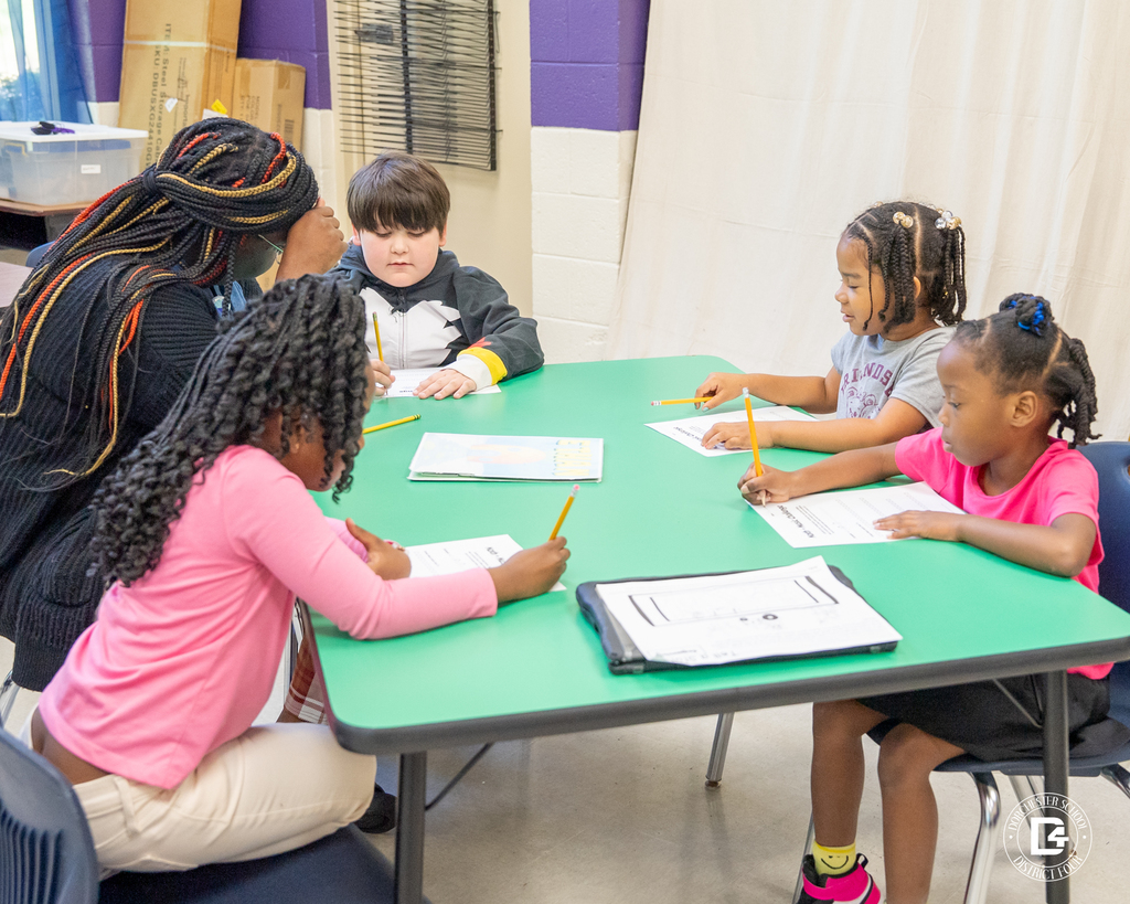A small group of students sits around a green table, working on their worksheets and talking through the challenge with a teacher.