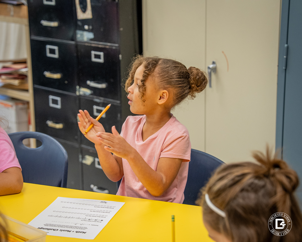 A young girl claps along with the rhythm while holding a pencil during Mr. Mansilla’s Math + Music Challenge. A worksheet sits on the yellow table in front of her.