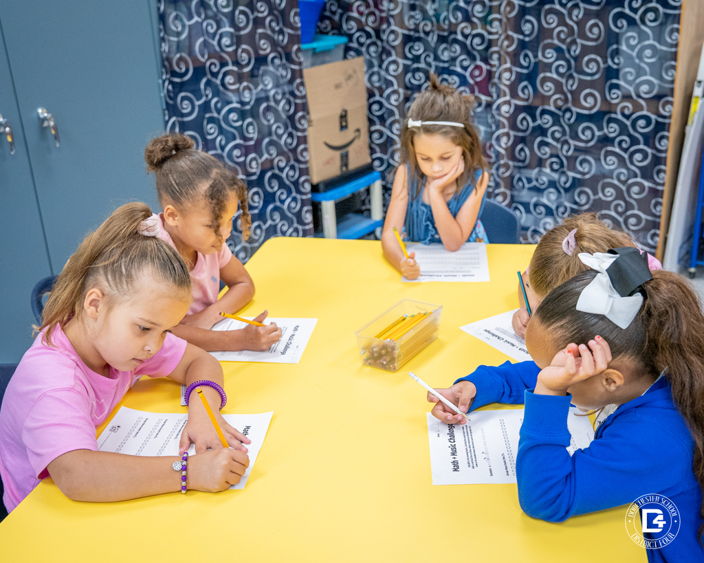 Several students gather around a yellow table completing their Math + Music Challenge worksheets as they listen for patterns in the music.