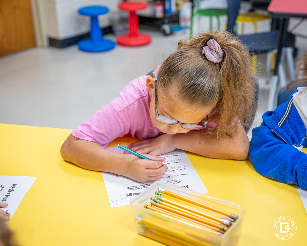 A student with glasses leans over her paper, writing carefully on the Math + Music Challenge worksheet at a yellow table.