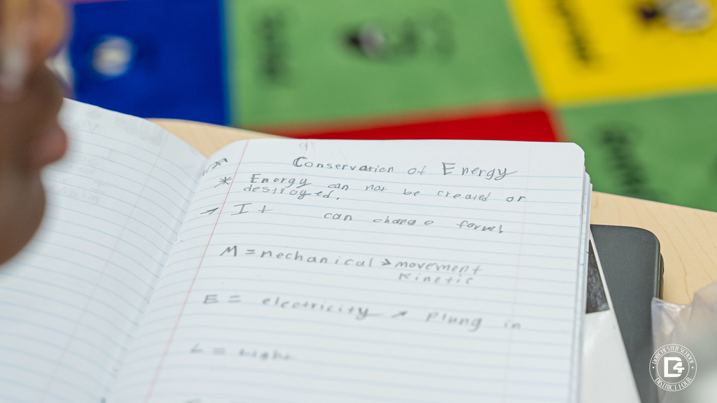 A student’s notebook open to notes labeled “Conservation of Energy,” describing how energy cannot be created or destroyed and can change form.