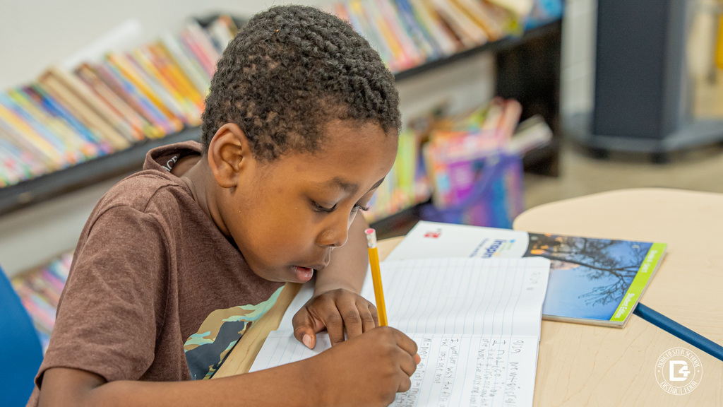 A student focuses on writing in his notebook during the energy discussion, surrounded by bookshelves in the background.