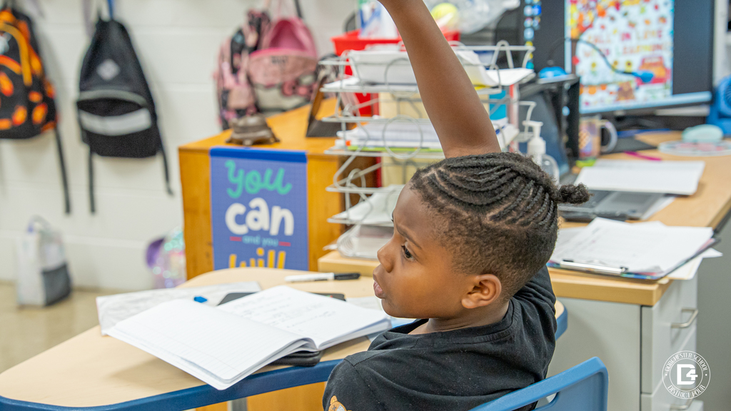 A student seated near the teacher’s desk raises his hand to participate in the discussion, with classroom posters and supplies in the background.