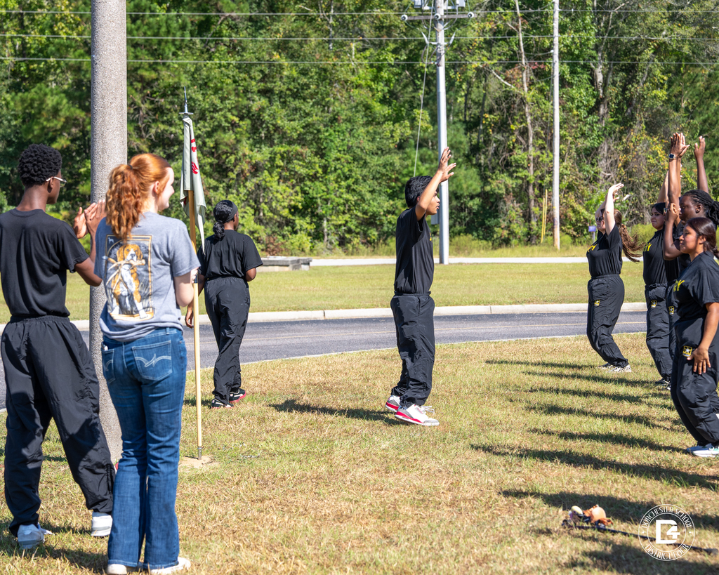 Cadets perform arm-raise exercises while instructors observe and encourage from the sidelines.
