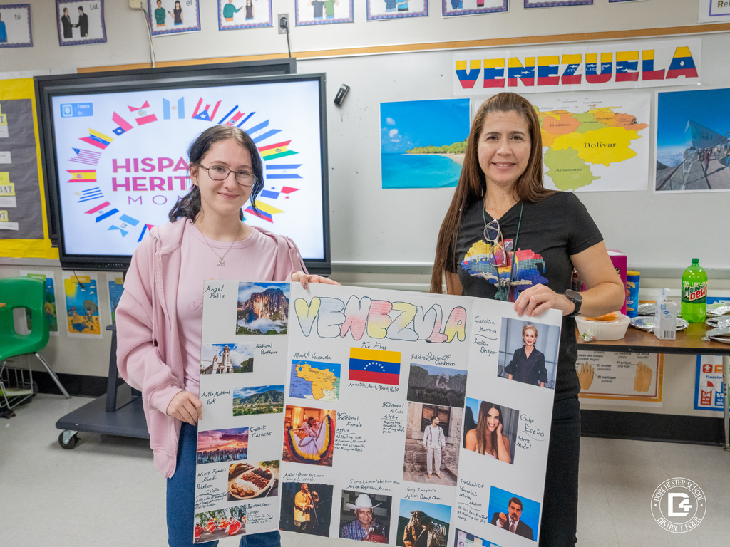A student and teacher smile while holding a colorful Venezuela project board in front of a digital display reading “Hispanic Heritage Month.”
