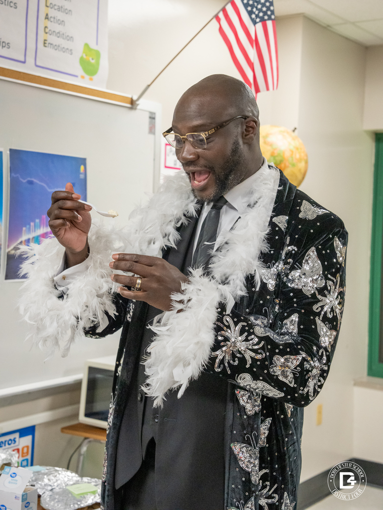 A male teacher dressed in a sparkling black and silver jacket with a white feather boa samples food while smiling during a classroom celebration.