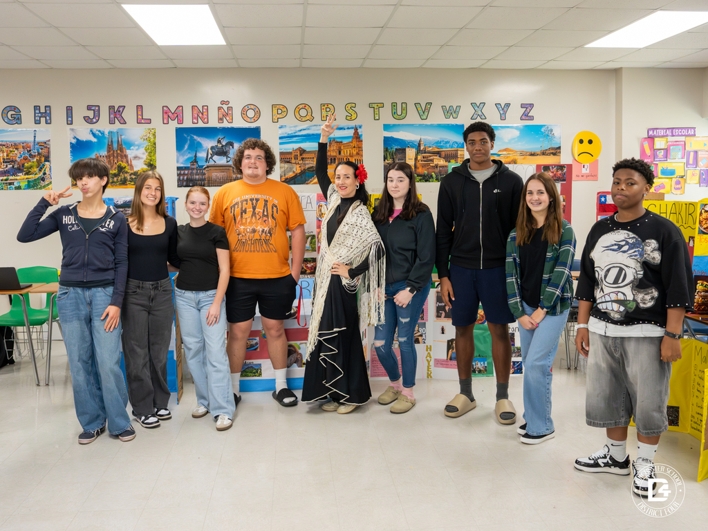 A group of high school students and their teacher pose together in a Spanish classroom decorated with cultural posters and alphabet letters.