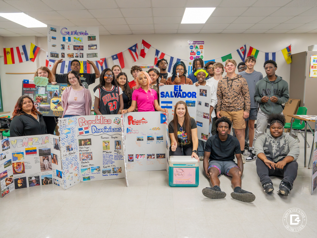 A large group of students poses with their Hispanic Heritage Month projects on countries including El Salvador, Venezuela, Puerto Rico, and the Dominican Republic, surrounded by international flags.