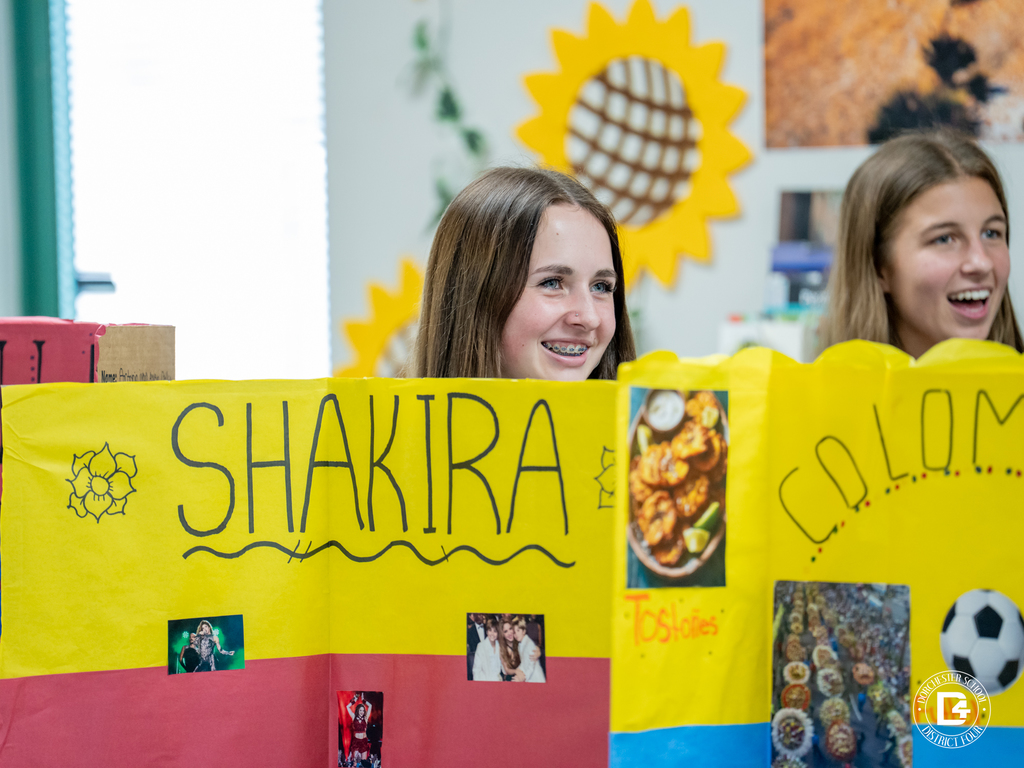 Two students stand behind a bright yellow and red project board about Shakira and Colombia, featuring pictures of the singer, Colombian food, and symbols.