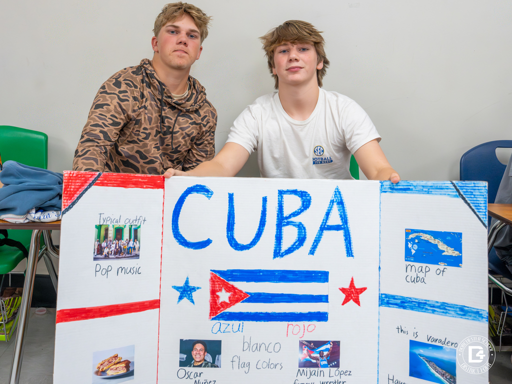Two students sit behind their trifold presentation board on Cuba, featuring the Cuban flag, maps, food, and famous people.