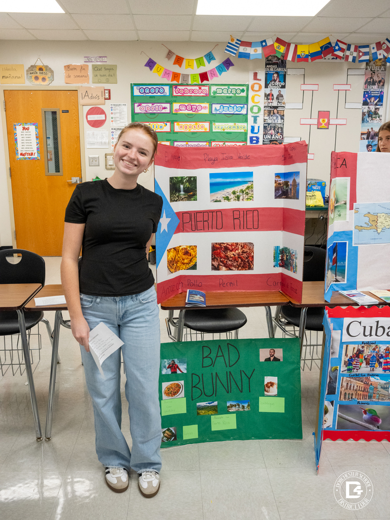 A smiling student stands beside a Puerto Rico project board decorated with photos, the flag, and images of Bad Bunny and Puerto Rican dishes.