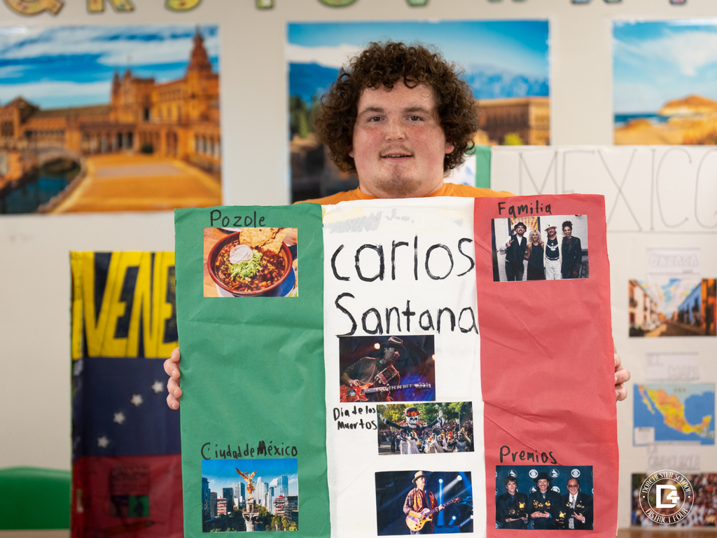 A student proudly displays a colorful trifold poster about Carlos Santana, featuring images of Mexican cuisine, family, and music awards.