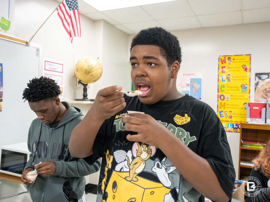 A student eats from a cup while participating in a classroom celebration, with another student standing beside him enjoying food.