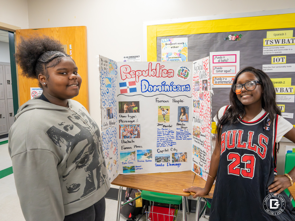 Two students stand beside their Dominican Republic project display featuring information on music, famous people, and traditional clothing.