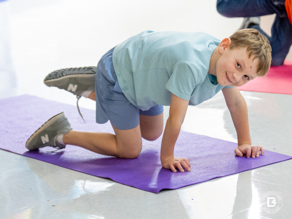 A smiling student in a light blue shirt performs a yoga pose on a purple mat during class, balancing on hands and knees while looking toward the camera.