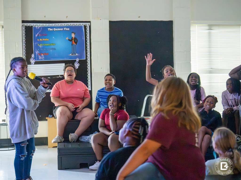 A group of students in a music classroom laughs and participates while one student sings into a yellow microphone. The atmosphere is lively and interactive.