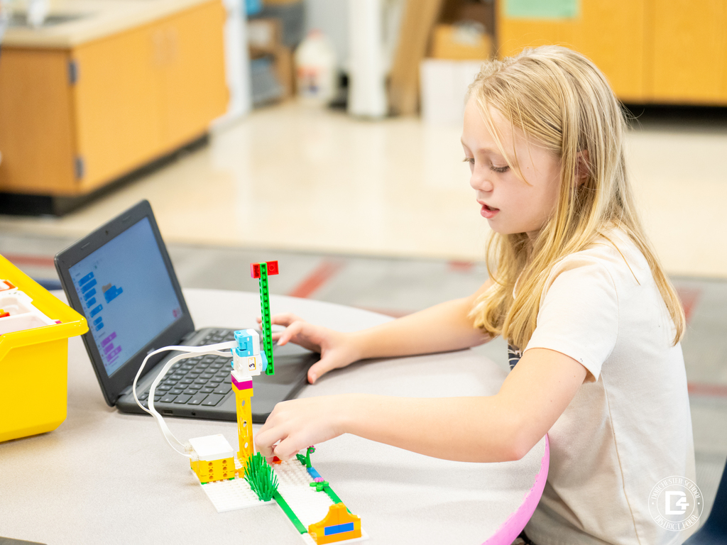 A blonde-haired student sits at a round table, programming a Lego robotics project on a laptop. A small motorized build is connected to the computer with a white cable.