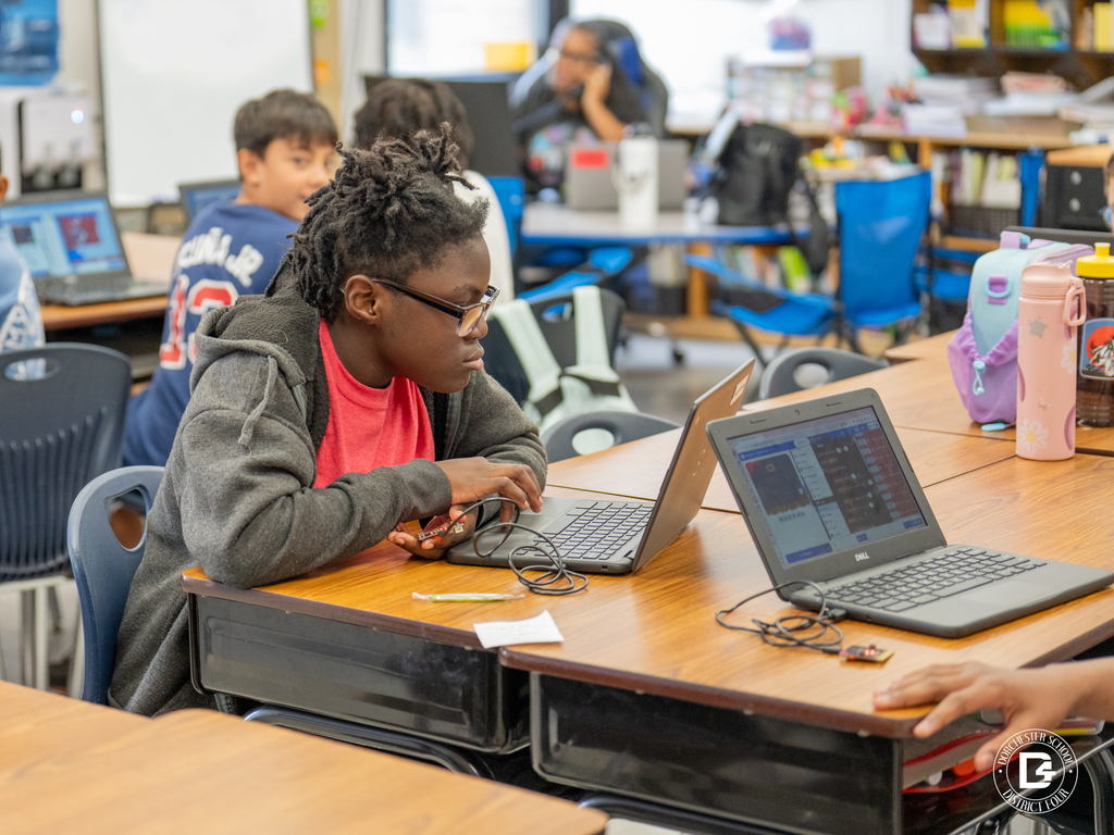 A student wearing glasses and a red shirt works intently on a coding project using a laptop in a classroom setting. Other students are visible in the background working on computers.