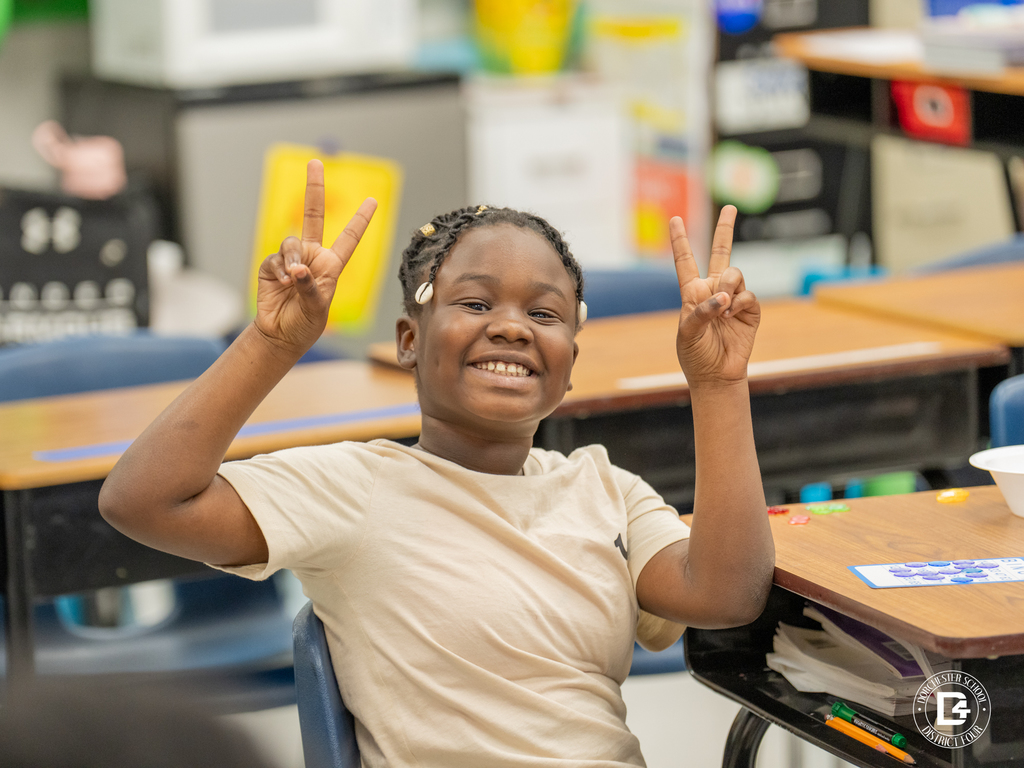 A cheerful student flashes double peace signs while sitting at a classroom desk. School supplies and other students are visible in the background.