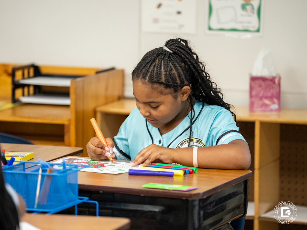 A young student with braided hair focuses on coloring a worksheet at her desk using a bright orange marker. School supplies and folders are on the desk, with classroom decor visible in the background.