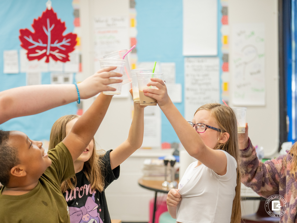 A group of students raises plastic cups in celebration inside a classroom. They are smiling and toasting together in front of colorful posters and classroom decor.
