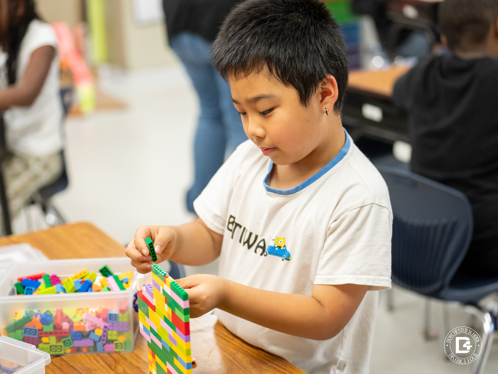 An elementary student builds a colorful Lego structure at his desk, carefully attaching pieces from a nearby container filled with building blocks.