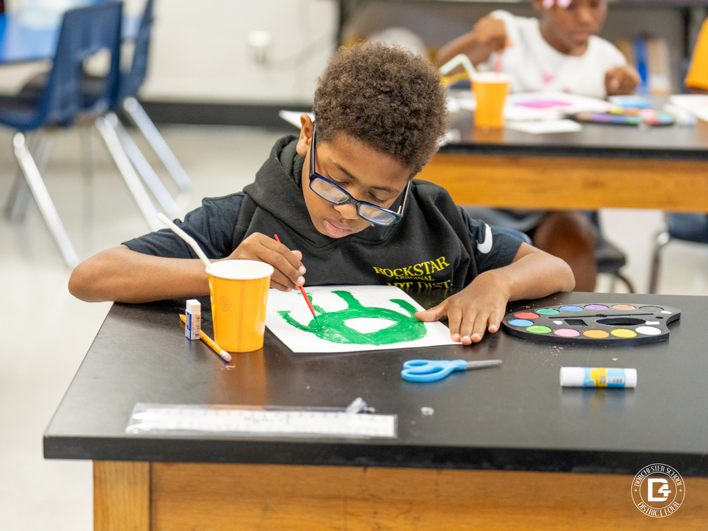 A student in glasses and a black hoodie paints a green figure on white paper during art class. Art supplies, scissors, and paint trays are spread across the table.