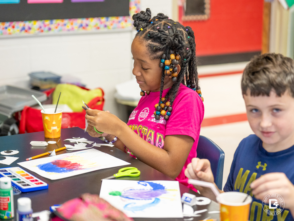 Two students work on colorful watercolor paintings at an art table, smiling and concentrating as they use scissors and brushes to complete their projects.