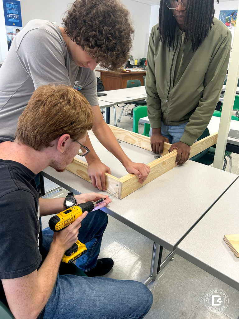 Three students assemble a wooden frame on a classroom table using a drill and teamwork.