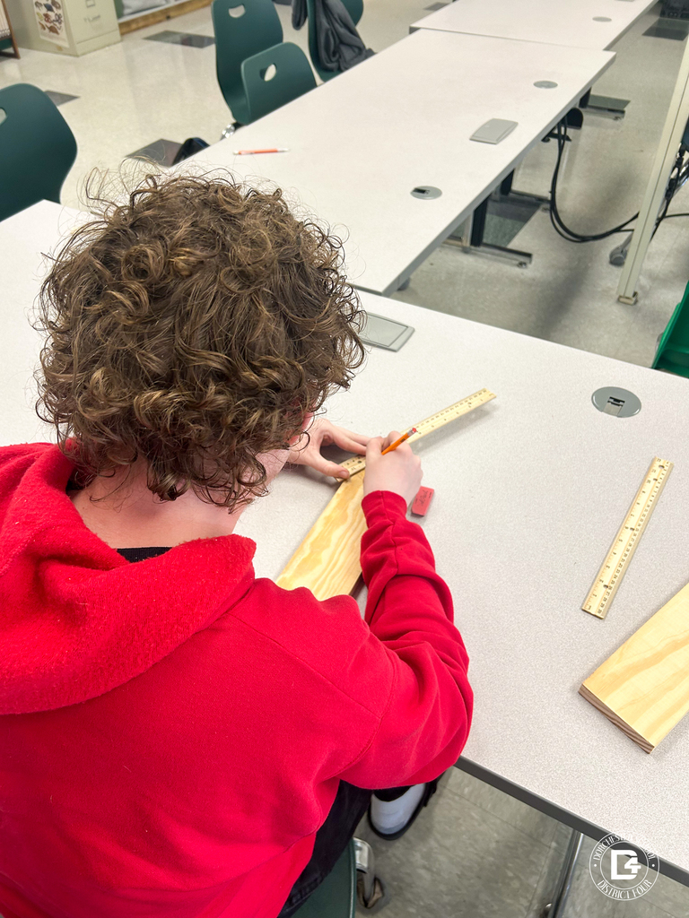 A student in a red hoodie measures and marks a piece of wood using a ruler at a classroom table.